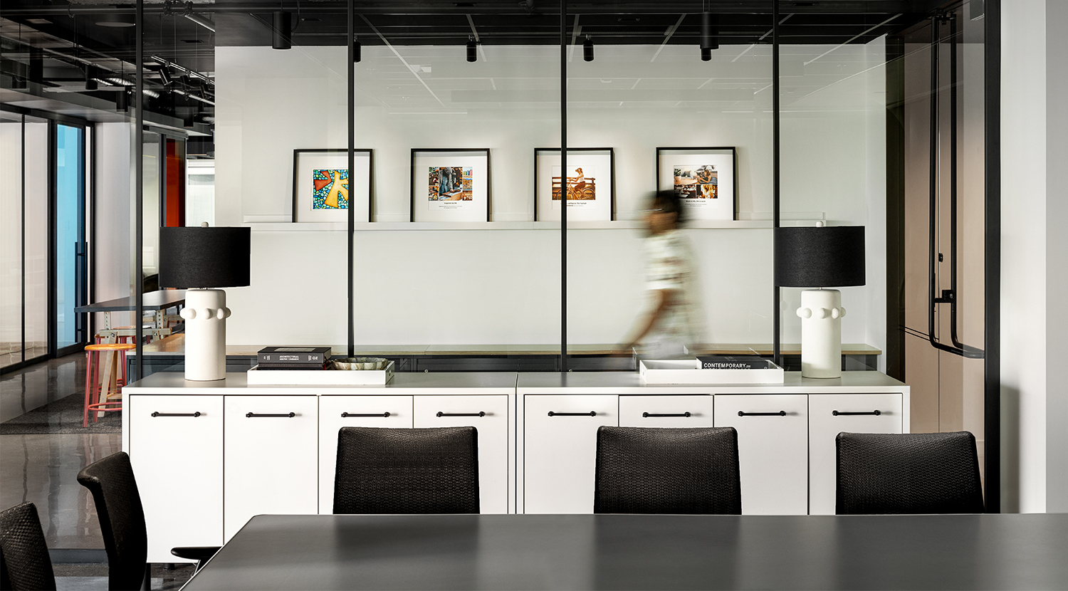 View rom inside glass wall boardroom, dark confrence table and black task chairs, row of white storage and contemporary lamps