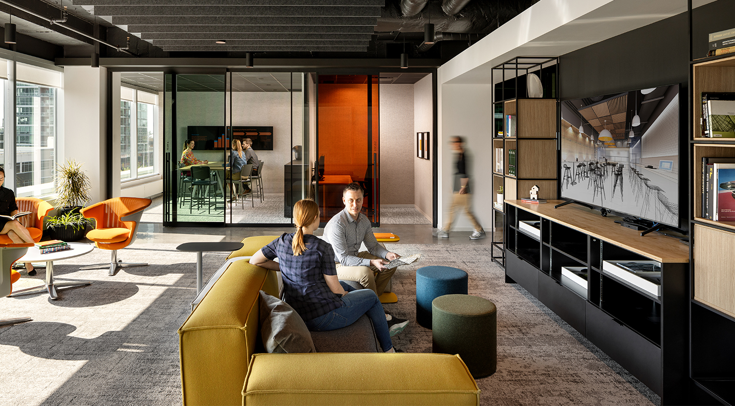 Yellow boxy sofa with blue and gray round ottomans in front of TV and bookcase, orange club chairs behind