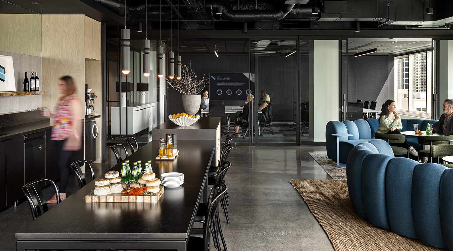 Busy work cafe with blue tufted sofa, round charcoal table and side chairs, backing onto glass wall boardroom
