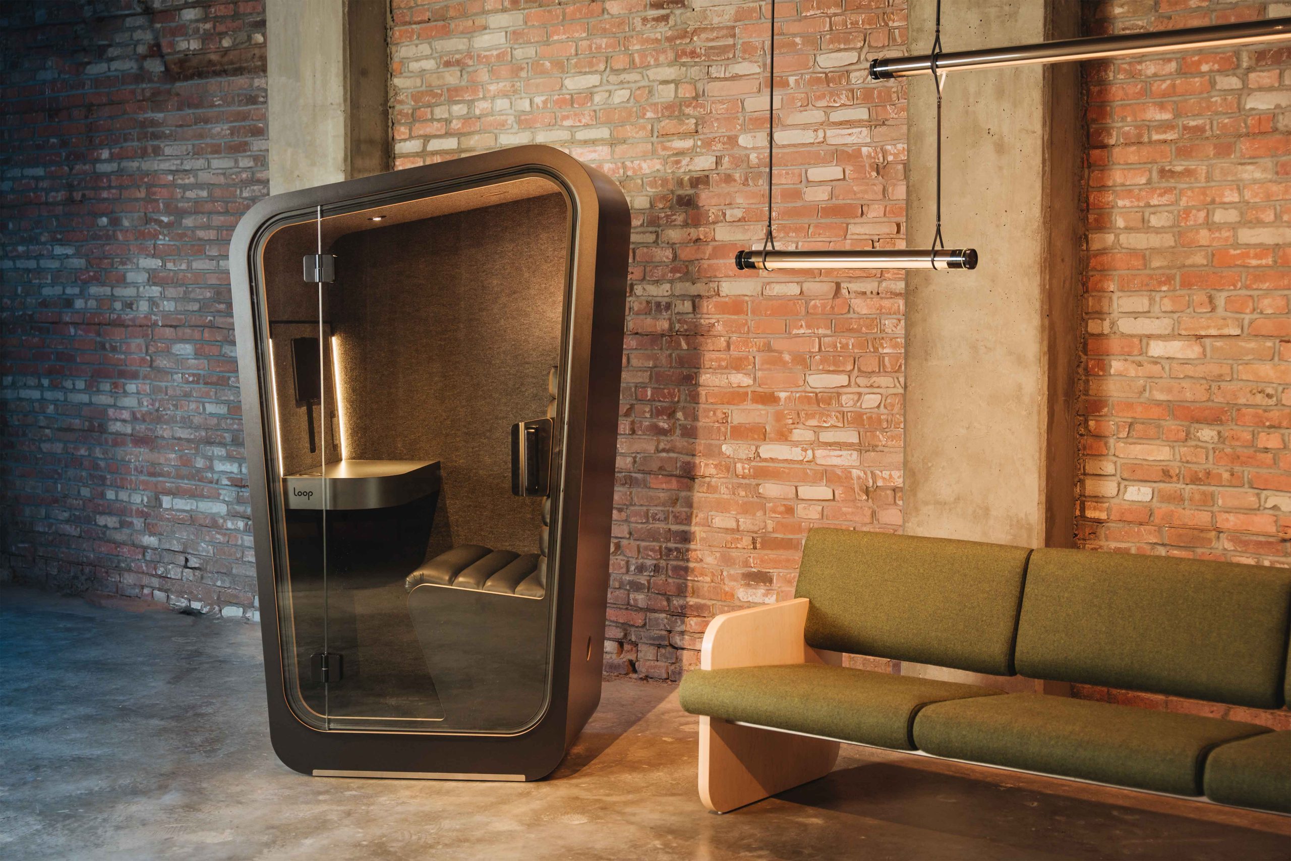 Solo Loop phone booth in black finishes in front of a brick wall with a green sofa