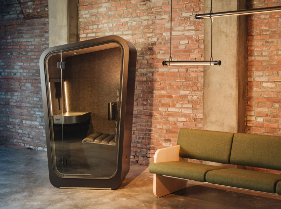 Solo Loop phone booth in black finishes in front of a brick wall with a green sofa